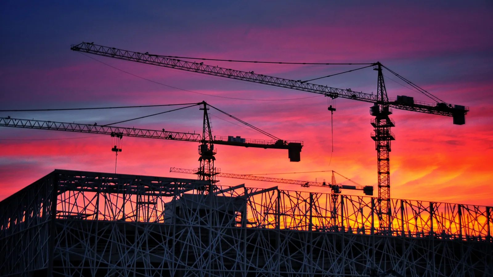 Construction cranes silhouetted against a pink and orange sunset